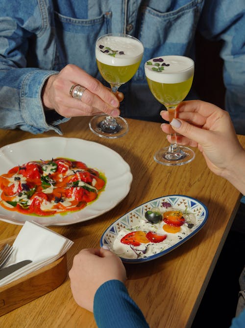 Man and Woman Clinking Cocktail Glasses over a Table with Food 