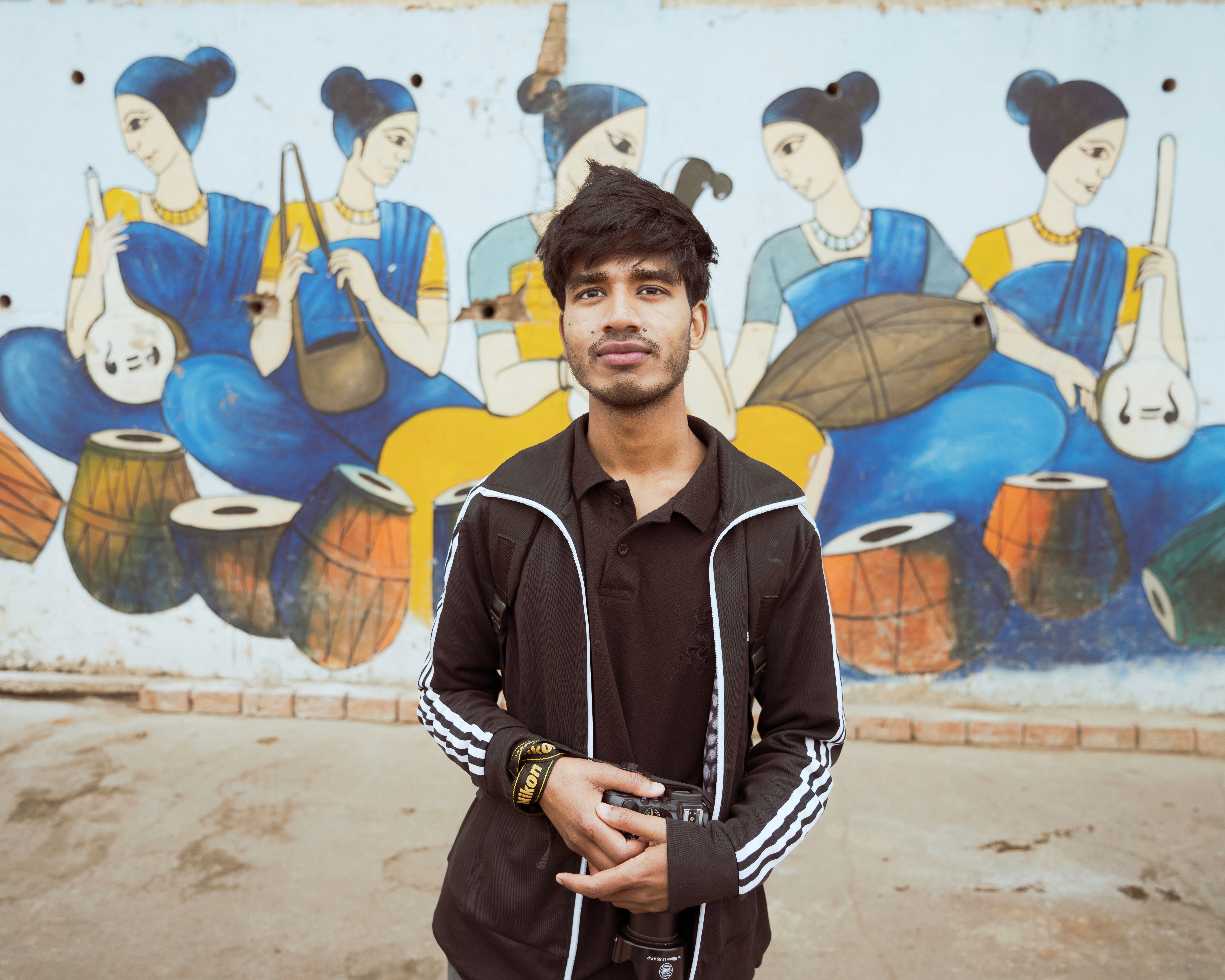 Portrait of a young man holding a camera standing in front of a vibrant mural depicting musicians.