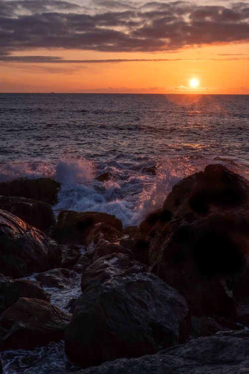 Free A breathtaking sunset view over the ocean with waves crashing against rocks, ideal for coastal themes. Stock Photo