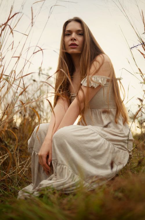 Free A long-haired woman in a white dress sits in a rural field during sunset, evoking a bohemian vibe. Stock Photo
