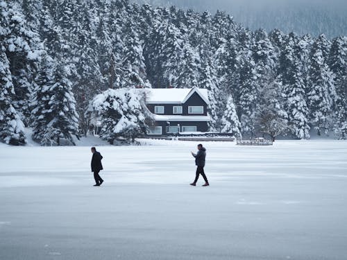 Two Men Walking on a Snowy Field near a House and a Forest 