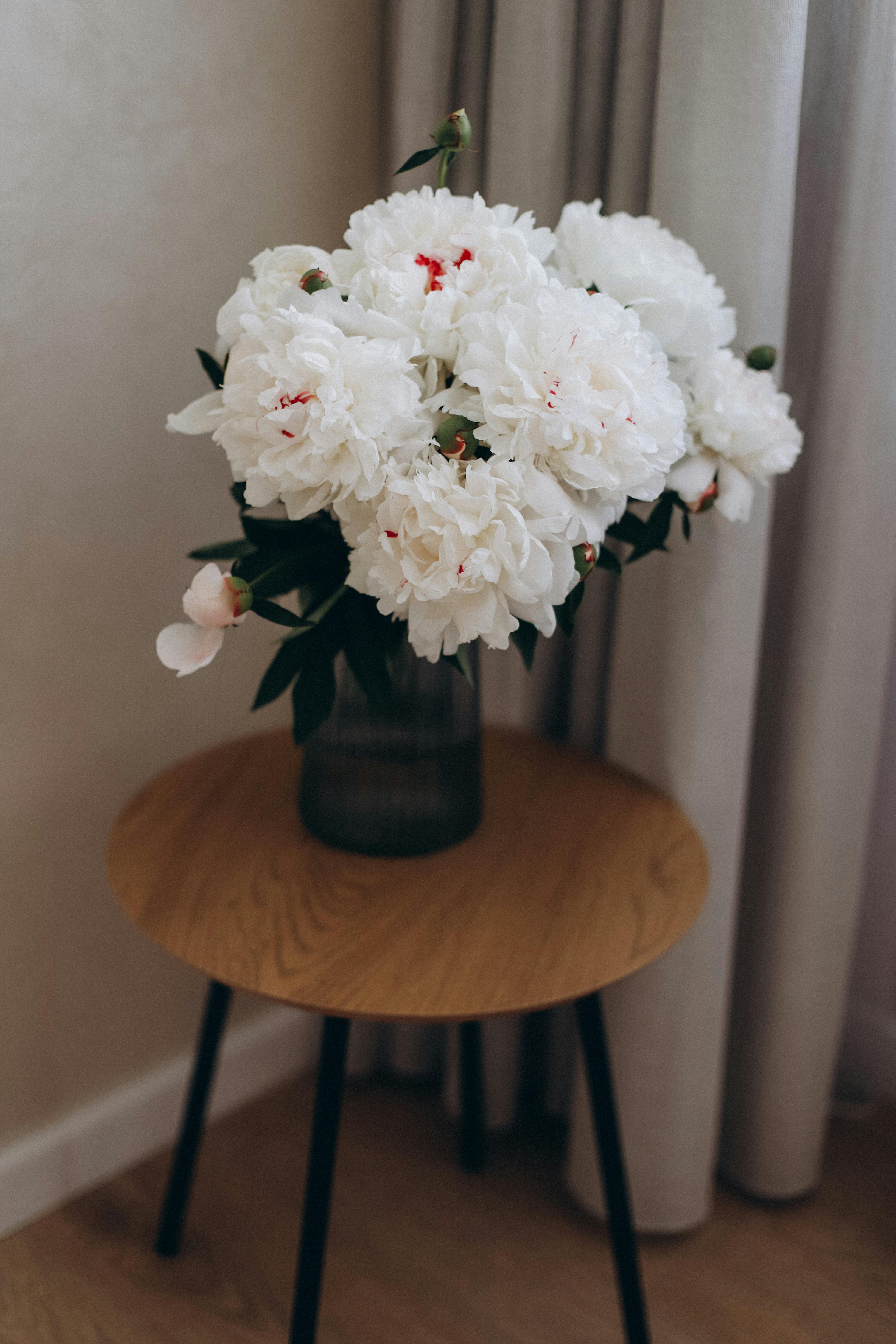 Beautiful bouquet of white peonies in a glass vase sitting on a wooden table indoors.