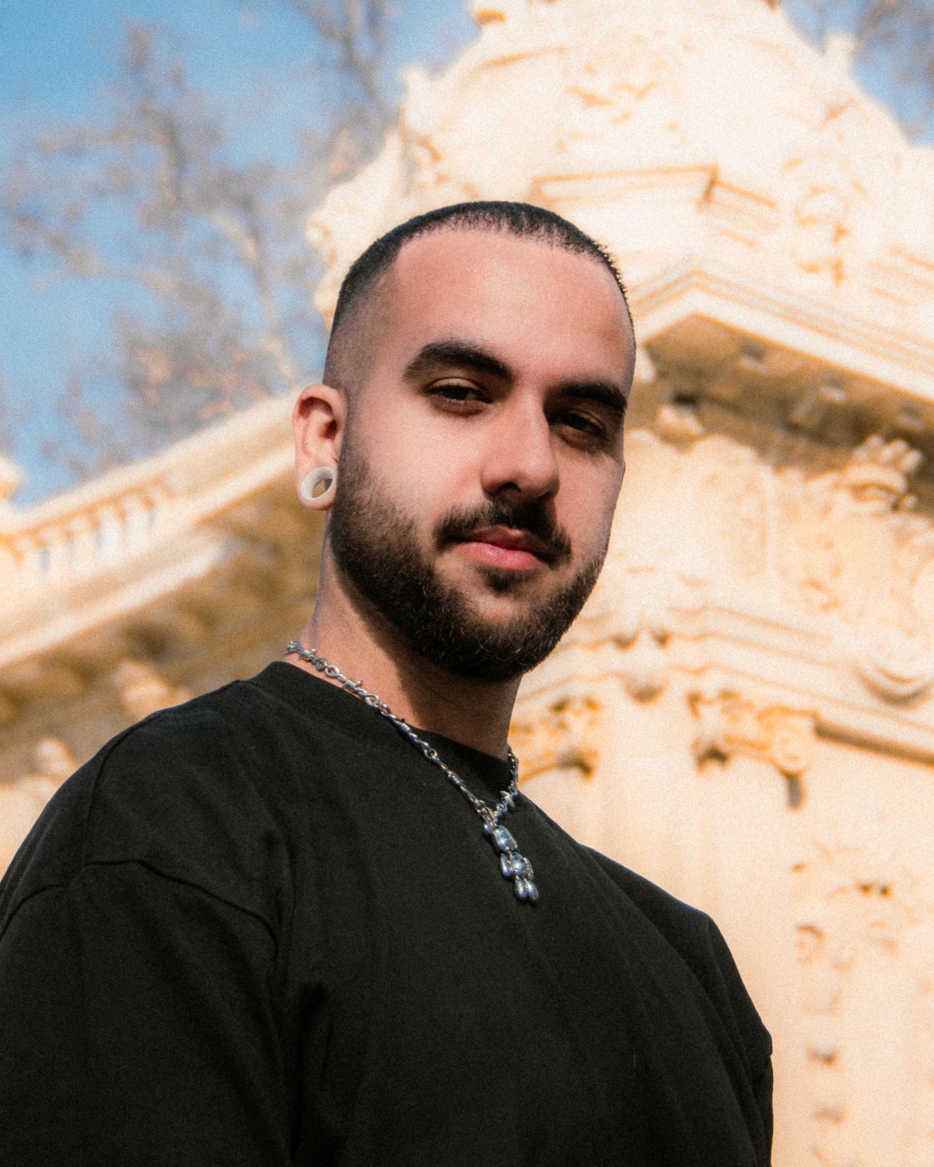 Close-up portrait of a man with ear tunnels and beard outdoors against ornate architecture.