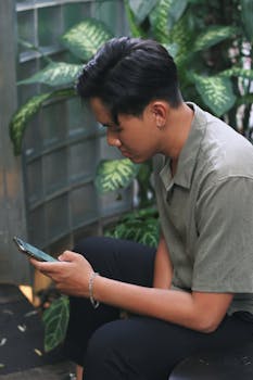 A young man in a casual outfit using a smartphone on a terrace surrounded by plants.