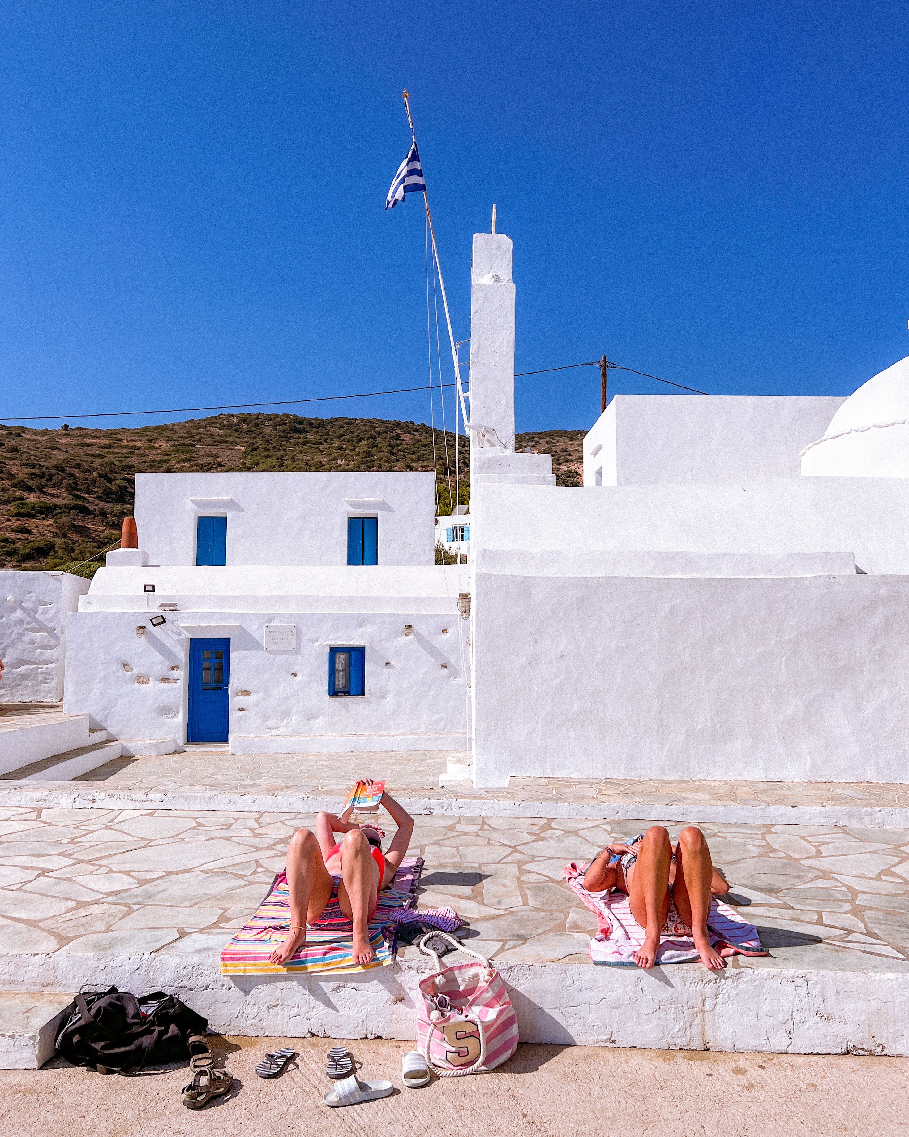 Two women sunbathing in front of traditional Greek architecture, showcasing vibrant summer vibes.