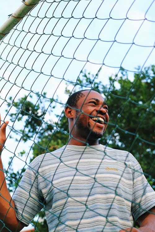 Low Angle Shot of a Young Man Standing behind a Chain Fence and Smiling 
