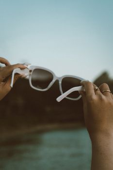 A pair of sunglasses held by hands against a blurred summer background, evoking travel vibes.