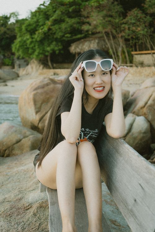Free Smiling Asian woman with sunglasses sitting on a beach bench, exuding summer vibes. Stock Photo