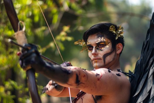 A man in fantasy makeup and costume aims a bow outdoors, embodying a warrior theme.