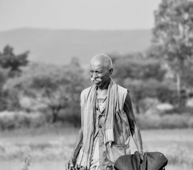 Black and white photo of an elderly man with a moustache walking through a rural area.