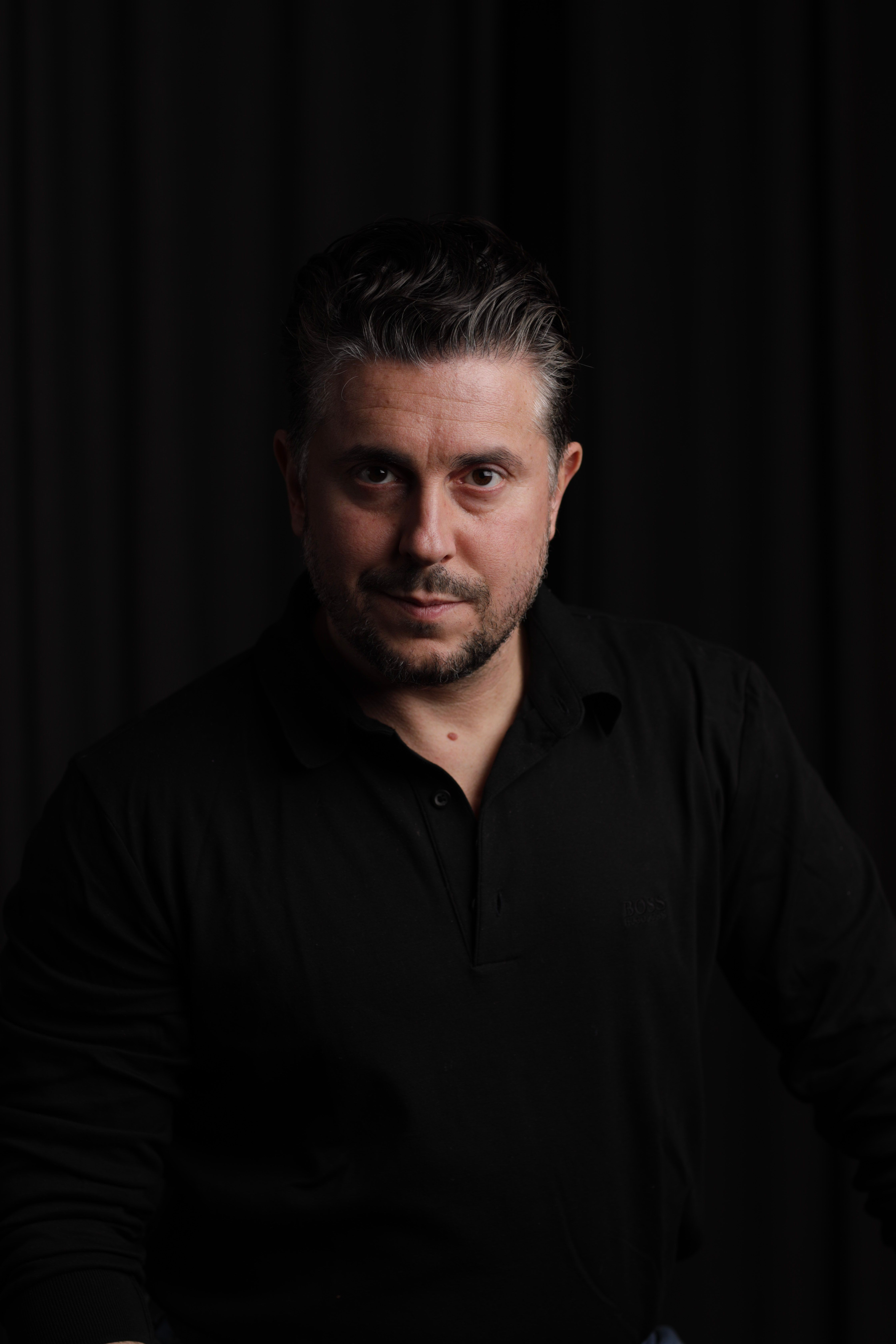 Portrait of a confident adult man with short hair in a black shirt, standing indoors.
