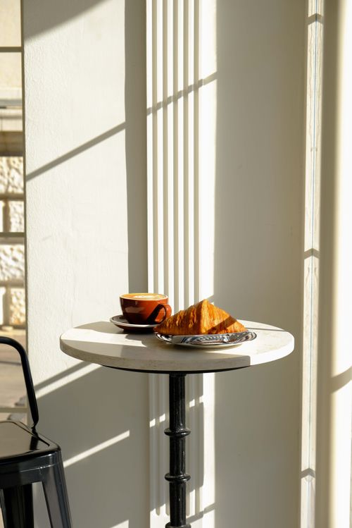 Free Warm sunlight illuminates a small cafe table with a croissant and coffee, capturing a serene breakfast moment. Stock Photo
