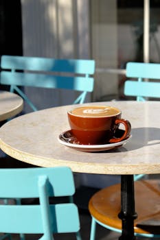 A brown cup of coffee with latte art on an outdoor cafe table with blue chairs.
