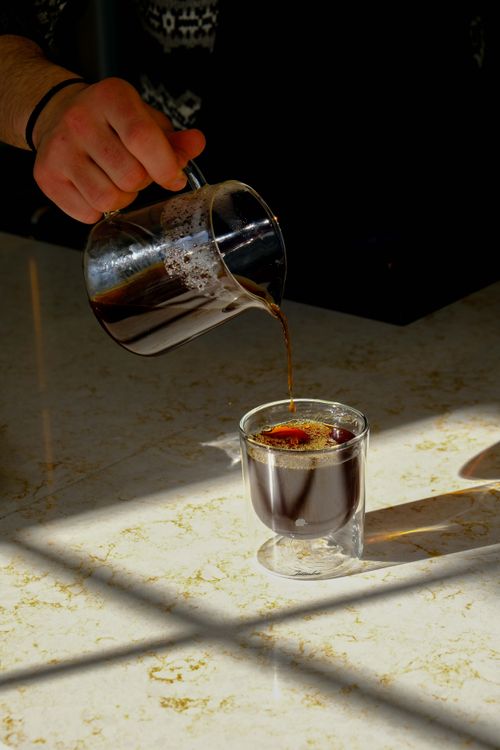 Free A person pouring freshly brewed coffee from a jug into a glass cup on a sunlit marble counter. Stock Photo