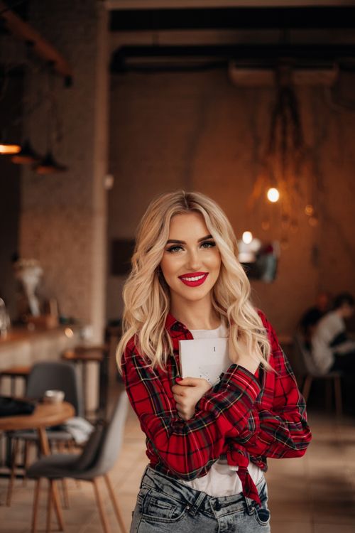 Free Cheerful blonde woman in a red plaid shirt smiling in a cozy cafe setting. Stock Photo