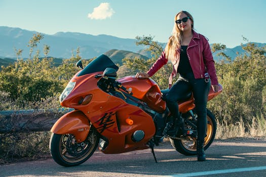 Fashionable woman in red jacket standing with motorcycle on scenic mountain road.