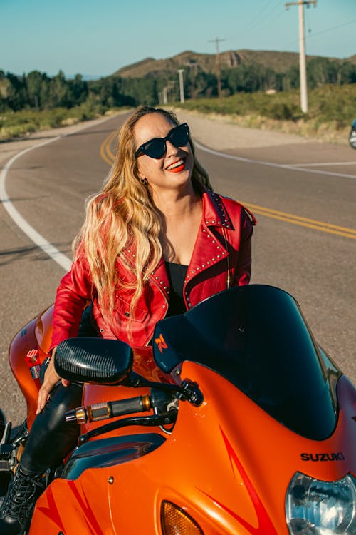 A woman in a leather jacket and sunglasses riding an orange motorcycle