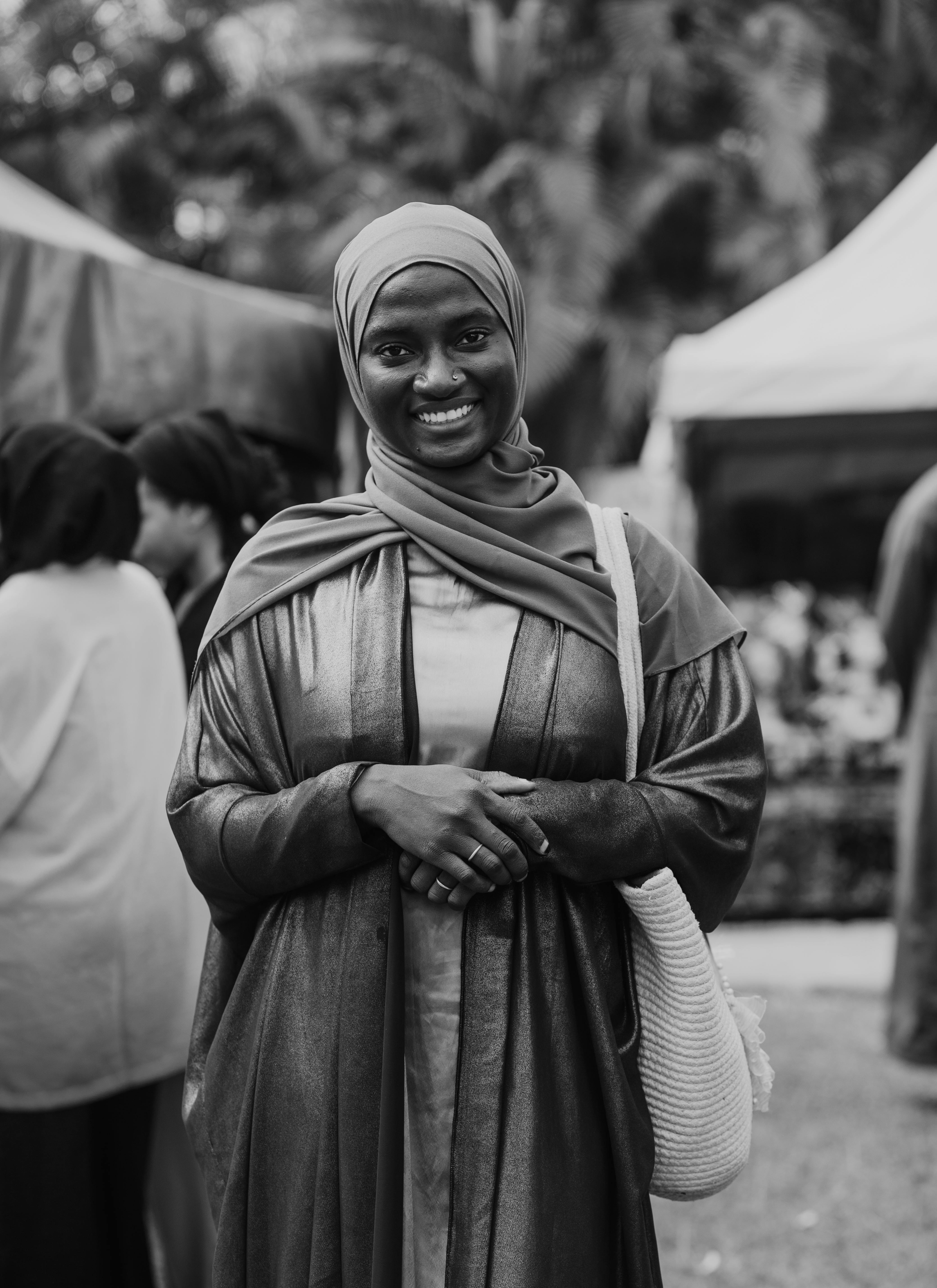 A smiling woman in a hijab and gown at an outdoor market in Abuja, Nigeria.