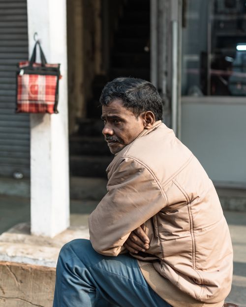 Free Side view of a man sitting on a city sidewalk, captured in an urban setting, wearing a winter jacket. Stock Photo