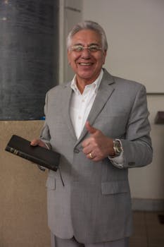 Happy senior man in a gray suit smiling and holding a book indoors, giving a thumbs up.