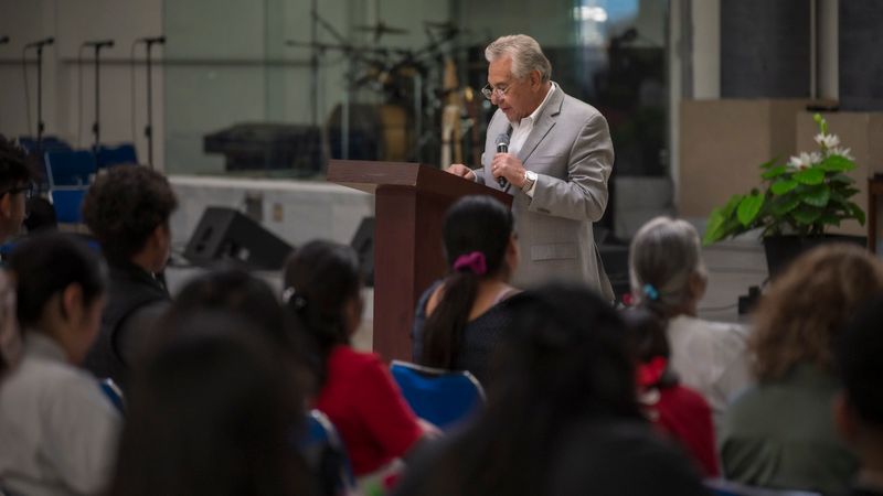 An elderly man in a gray suit delivers a speech to a seated audience indoors.