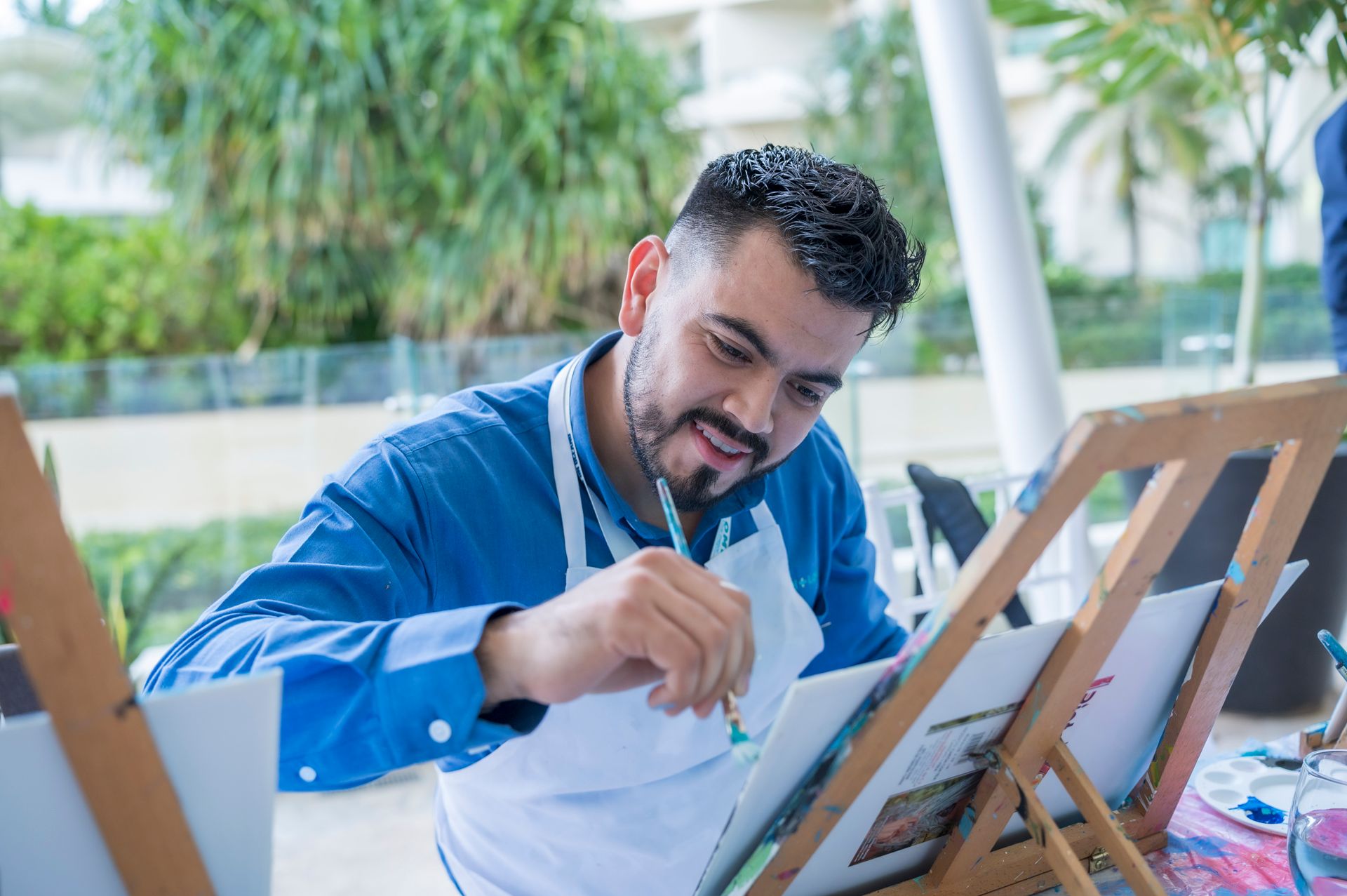 A man wearing a blue shirt and apron happily paints outdoors during a workshop.
