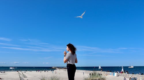A woman standing on the beach with a seagull flying above her