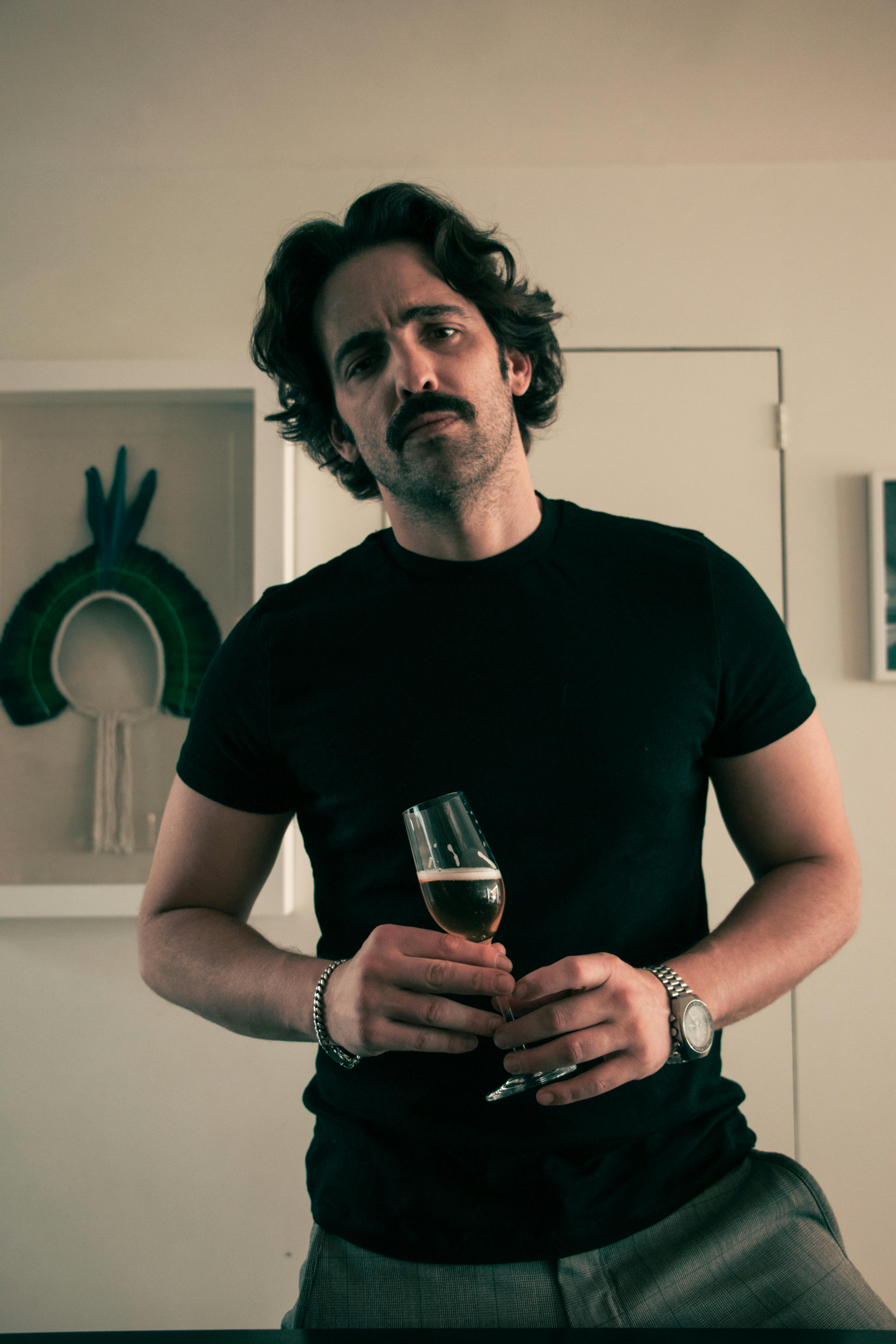Adult man with mustache holding a drink in a casual indoor setting, wearing a black t-shirt.