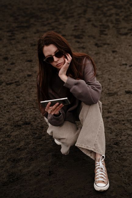 A young woman with sunglasses and casual attire using a smartphone on a sandy beach, captured in a moody tone.