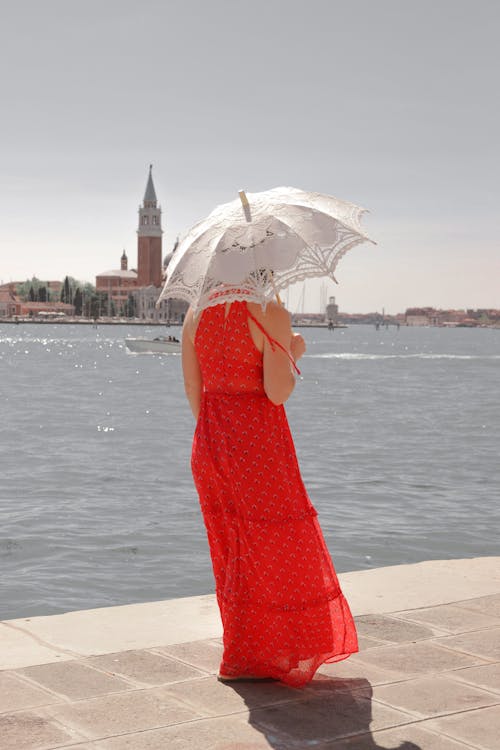 Back View of a Woman in a Red Dress Holding a Sun Umbrella Walking on the Shore in Venice, Italy 