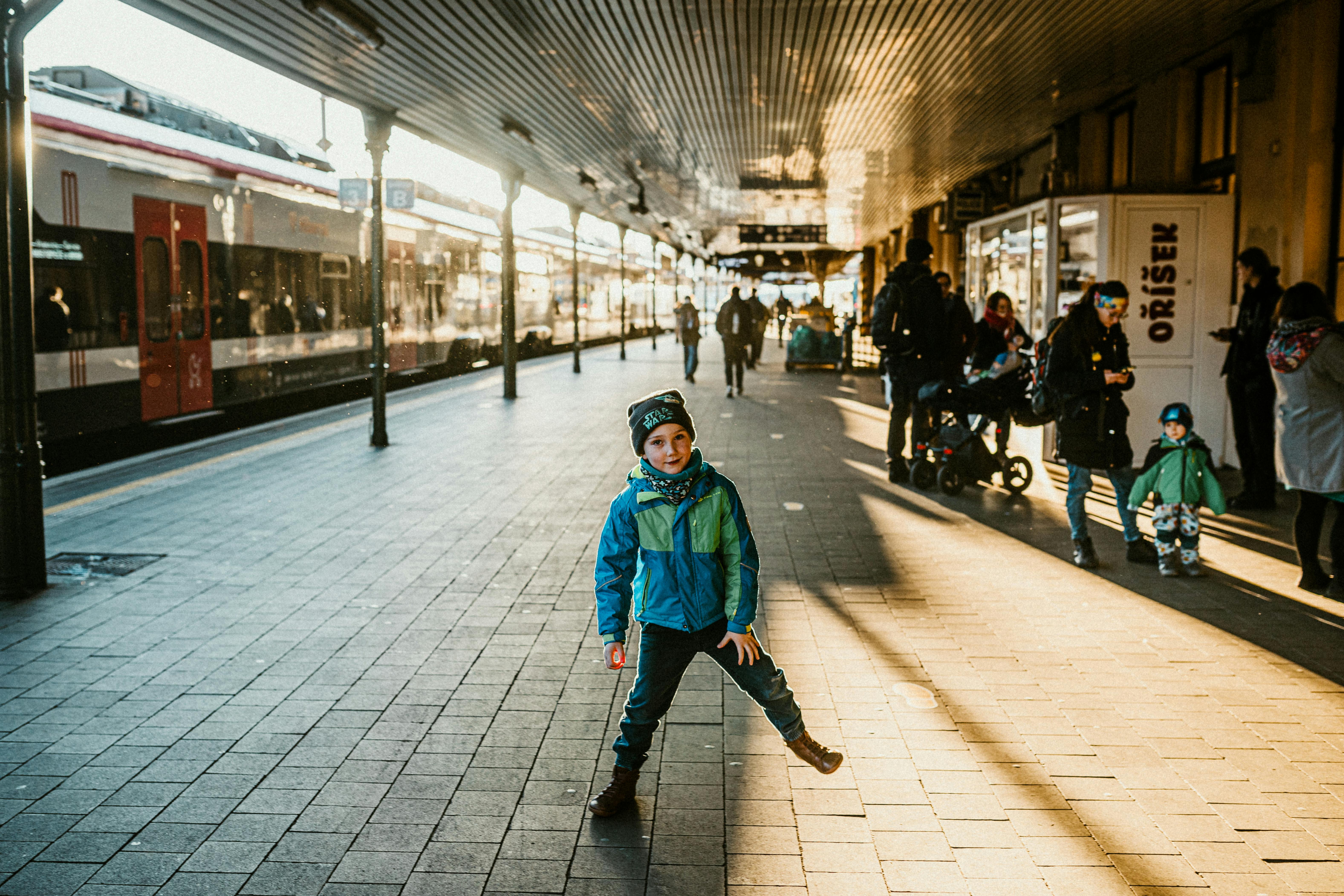 A young boy in a vibrant jacket plays joyfully at a bustling urban railway station.