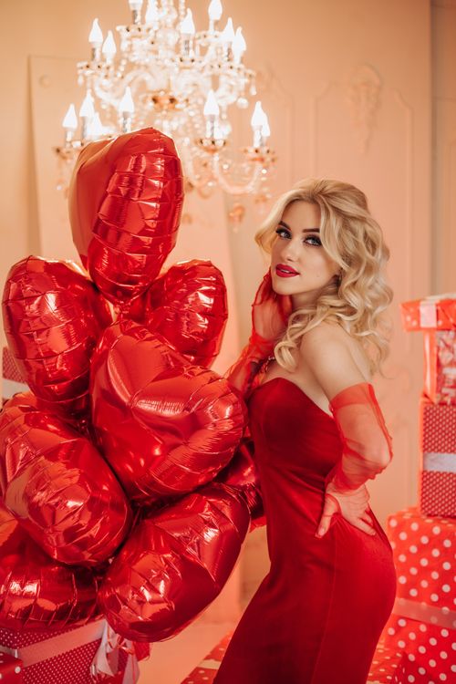 Free Blonde woman in a red dress poses with heart-shaped balloons in an elegant setting. Stock Photo