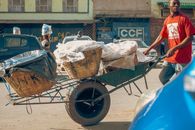 A man in red shirt pushes a wheelbarrow with baskets on a bustling Lusaka street.