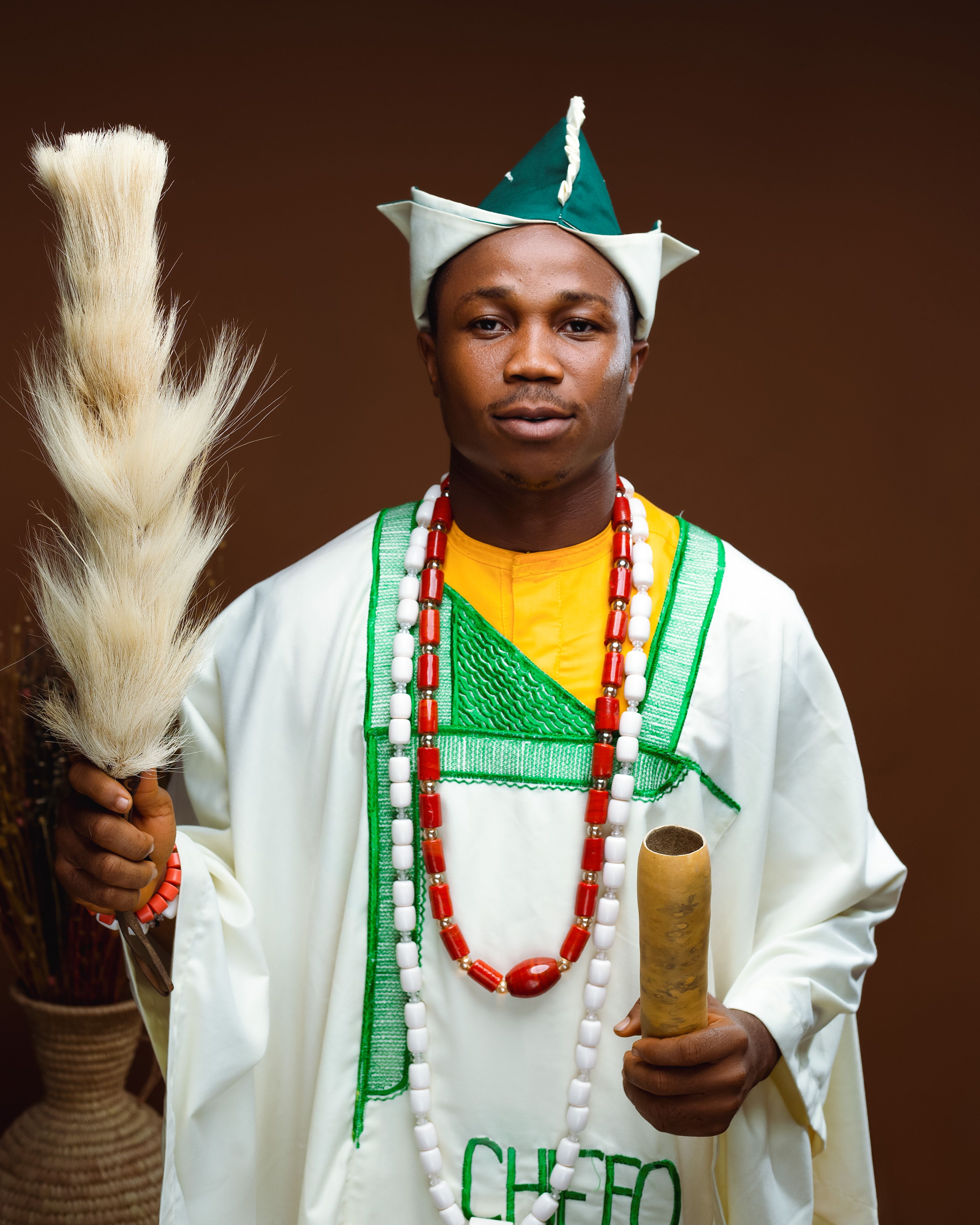 African man in traditional Nigerian clothing holding cultural symbols in studio setting.
