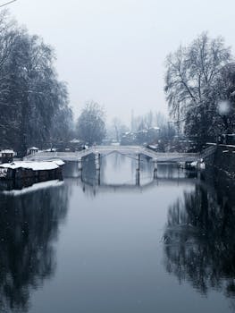Peaceful winter scene with a snow-covered bridge and calm river reflecting the surrounding landscape.
