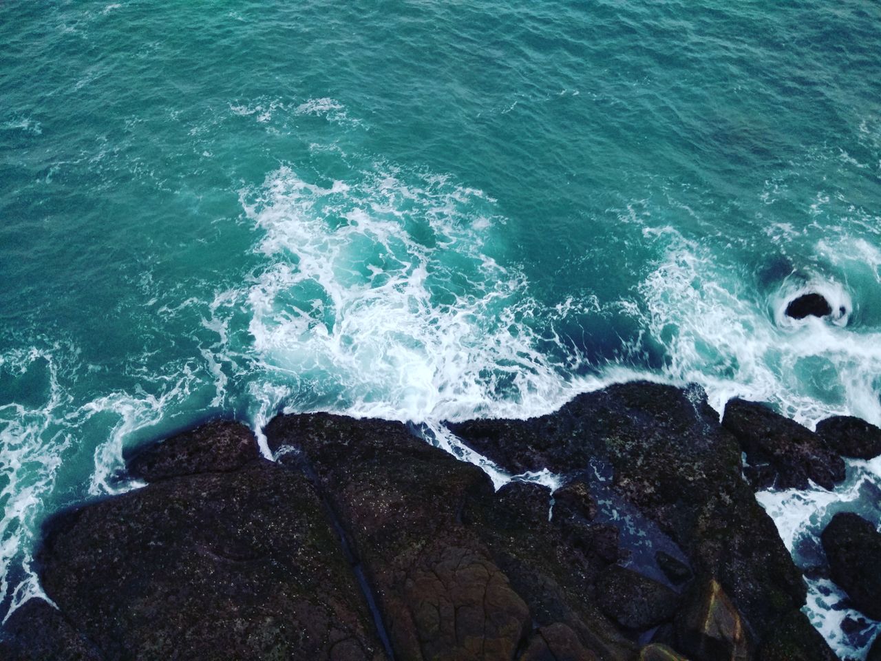 aerial photograph of turquoise ocean waves crashing against a rocky sh