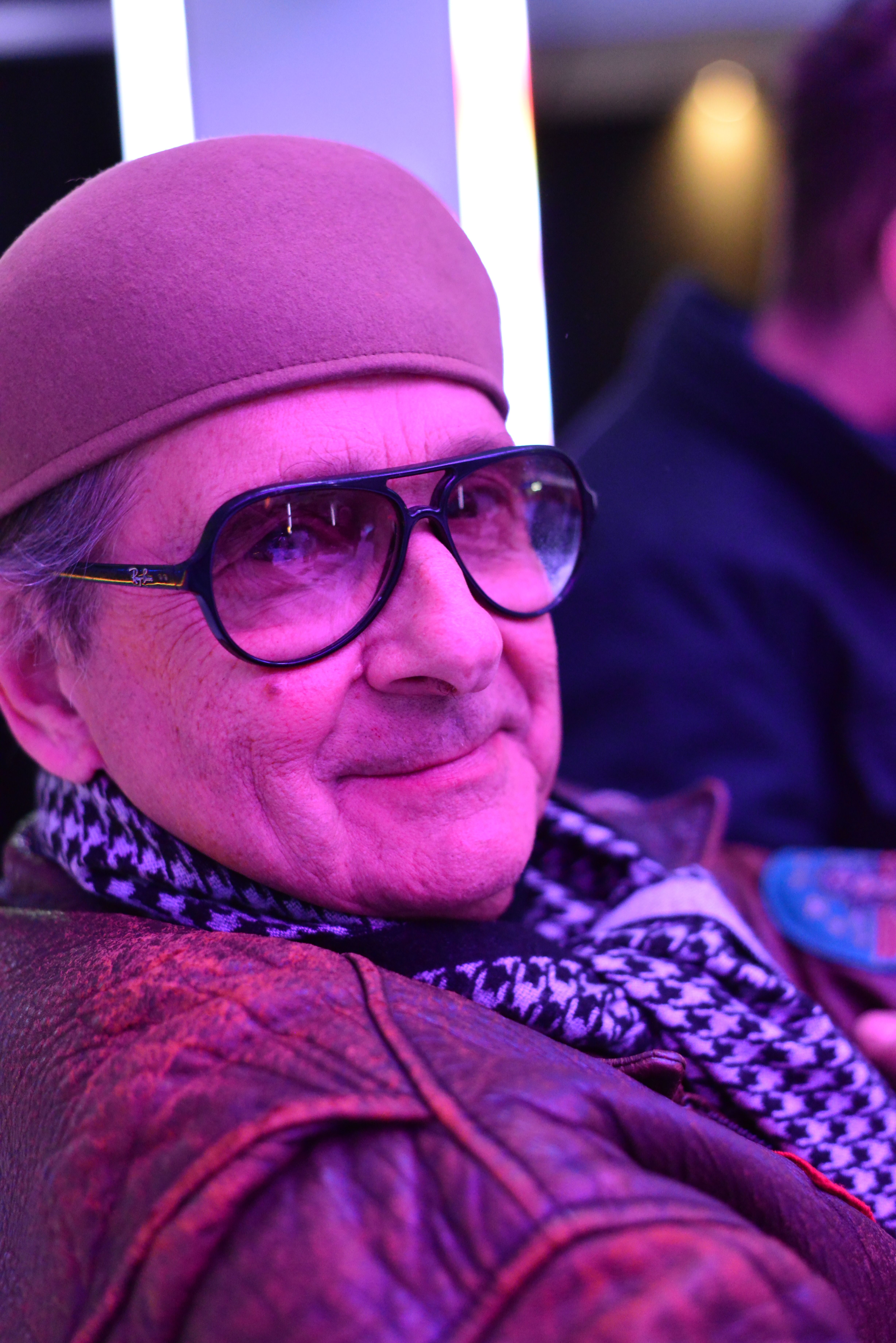 Close-up portrait of an elderly man wearing sunglasses and a hat under pink lighting.