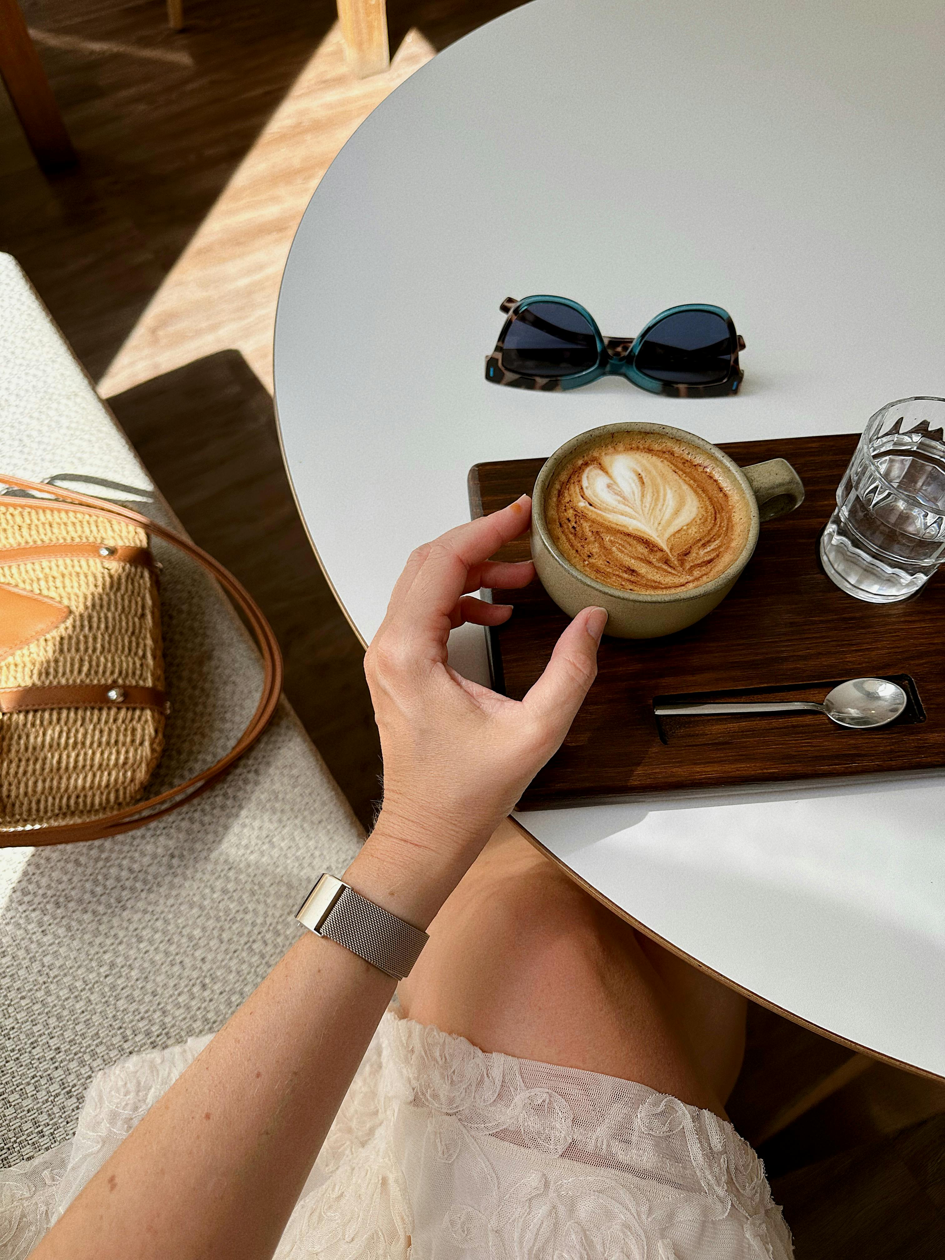 A woman enjoying a latte at a café table, featuring sunglasses and a woven bag.