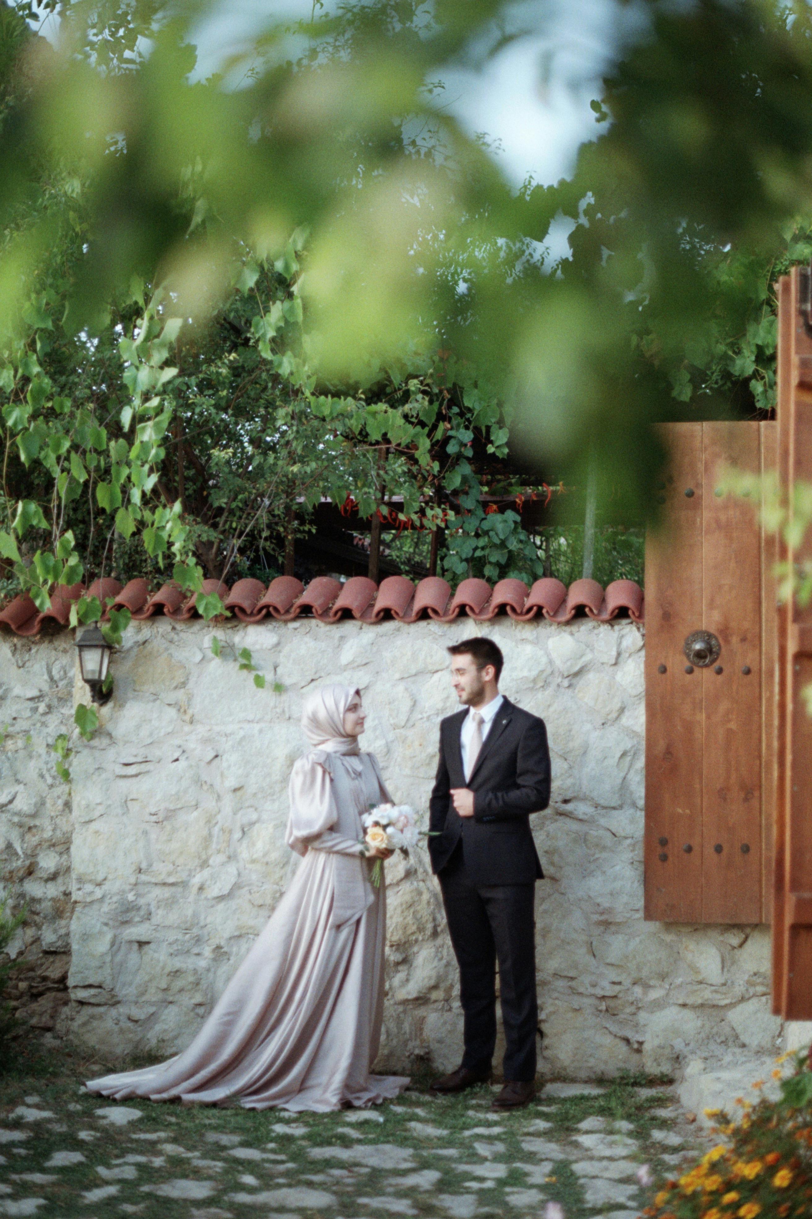 A beautifully dressed couple stands by a rustic wall in an outdoor setting, embodying elegance.