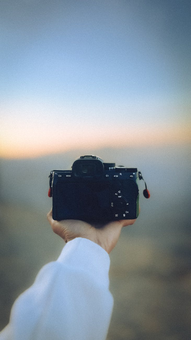 A person holds a camera against a blurred sunset backdrop, capturing the serene moment.