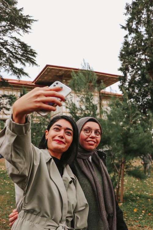 Free Two women smiling and taking a selfie outdoors with a smartphone in front of a building. Stock Photo