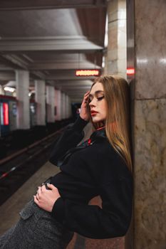 Fashionable woman with long hair posing against a wall in a subway station, showcasing urban style.