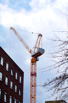 A tall construction crane beside a modern urban building under a cloudy sky.
