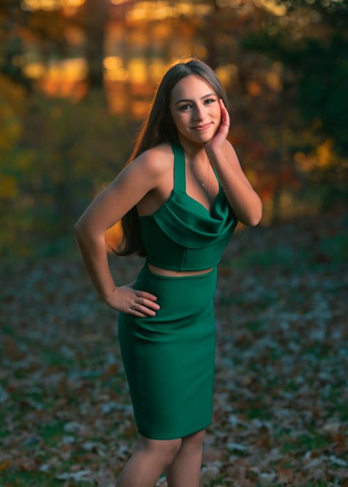Free A young woman poses elegantly in a green dress amidst a forest at sunset, with vibrant autumn leaves. Stock Photo