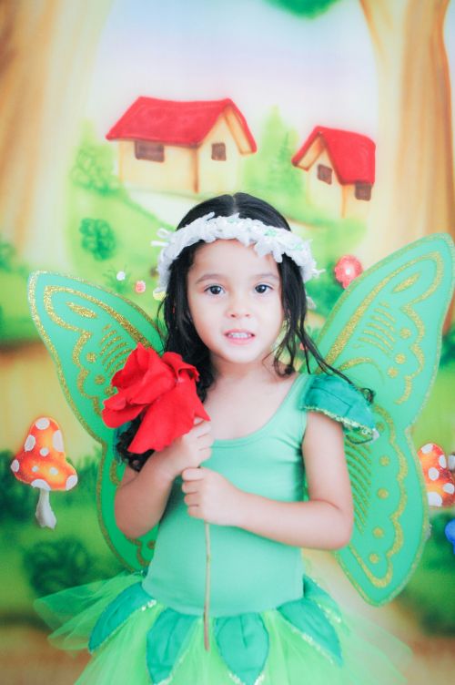 Free Adorable child in green fairy costume with wings holding red flower, against whimsical backdrop. Stock Photo