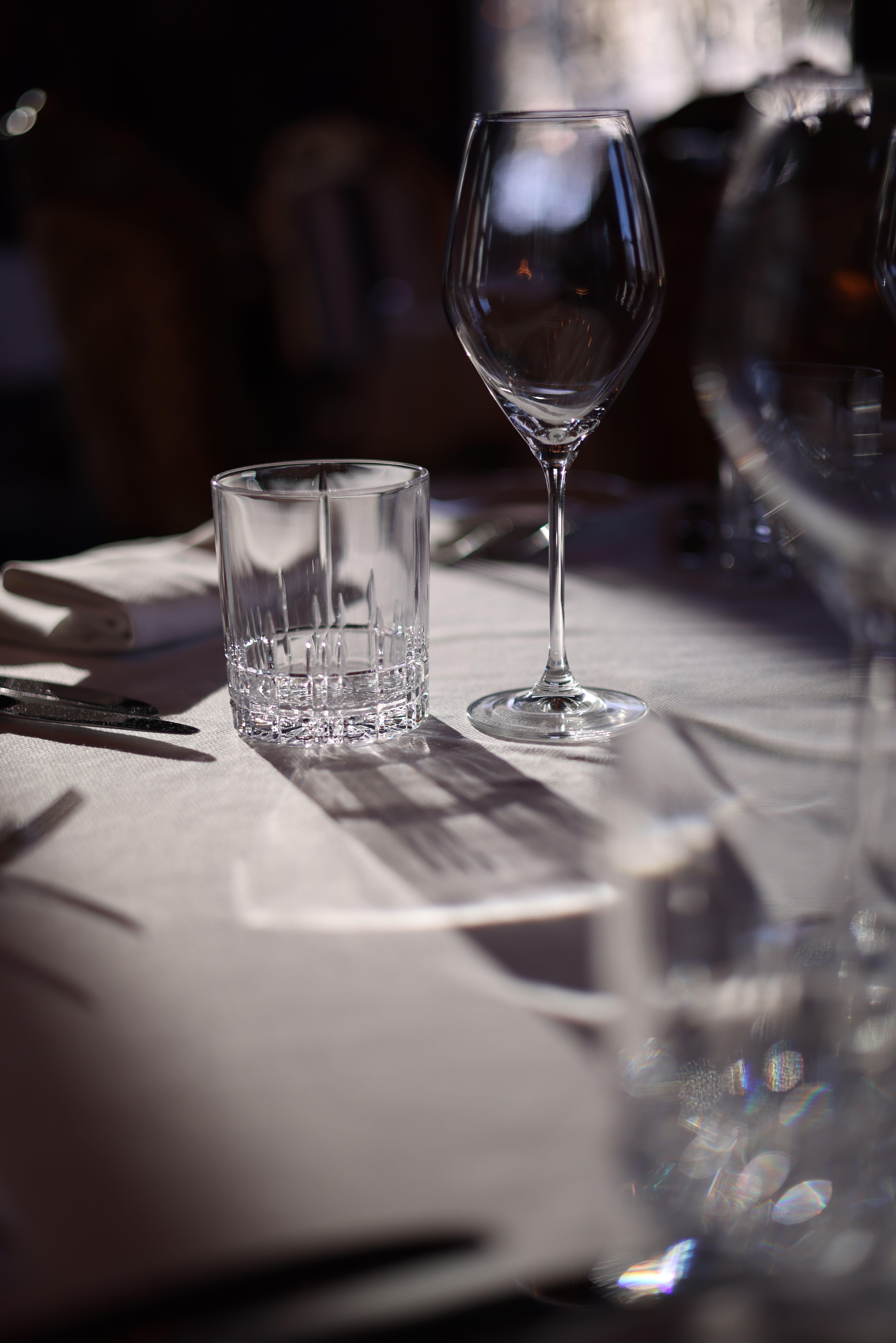 Close-up of wineglass and tumbler on a sunlit dining table, soft focus.
