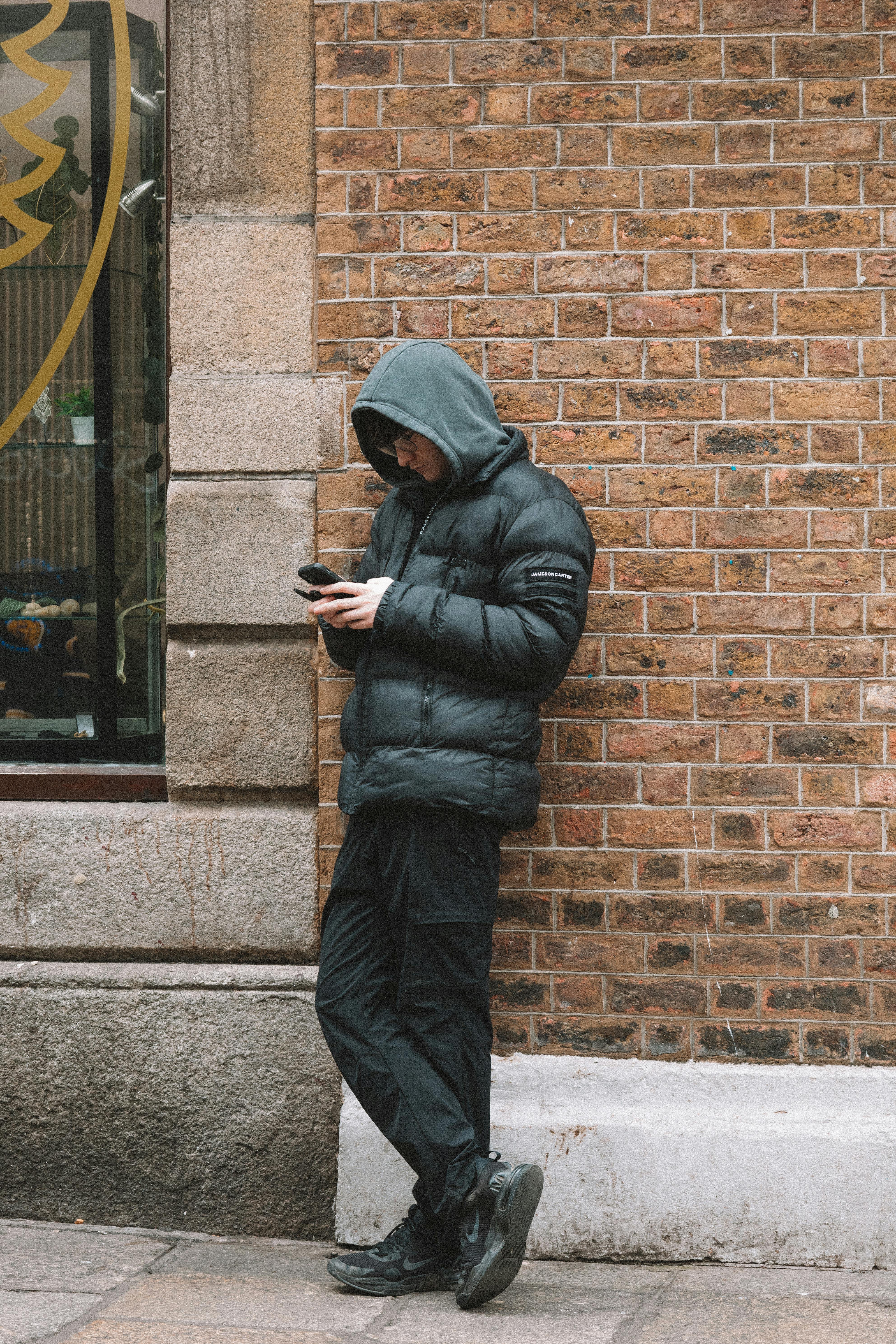 Free A young man in a puffer jacket uses his phone while leaning against a brick wall in an urban area. Stock Photo