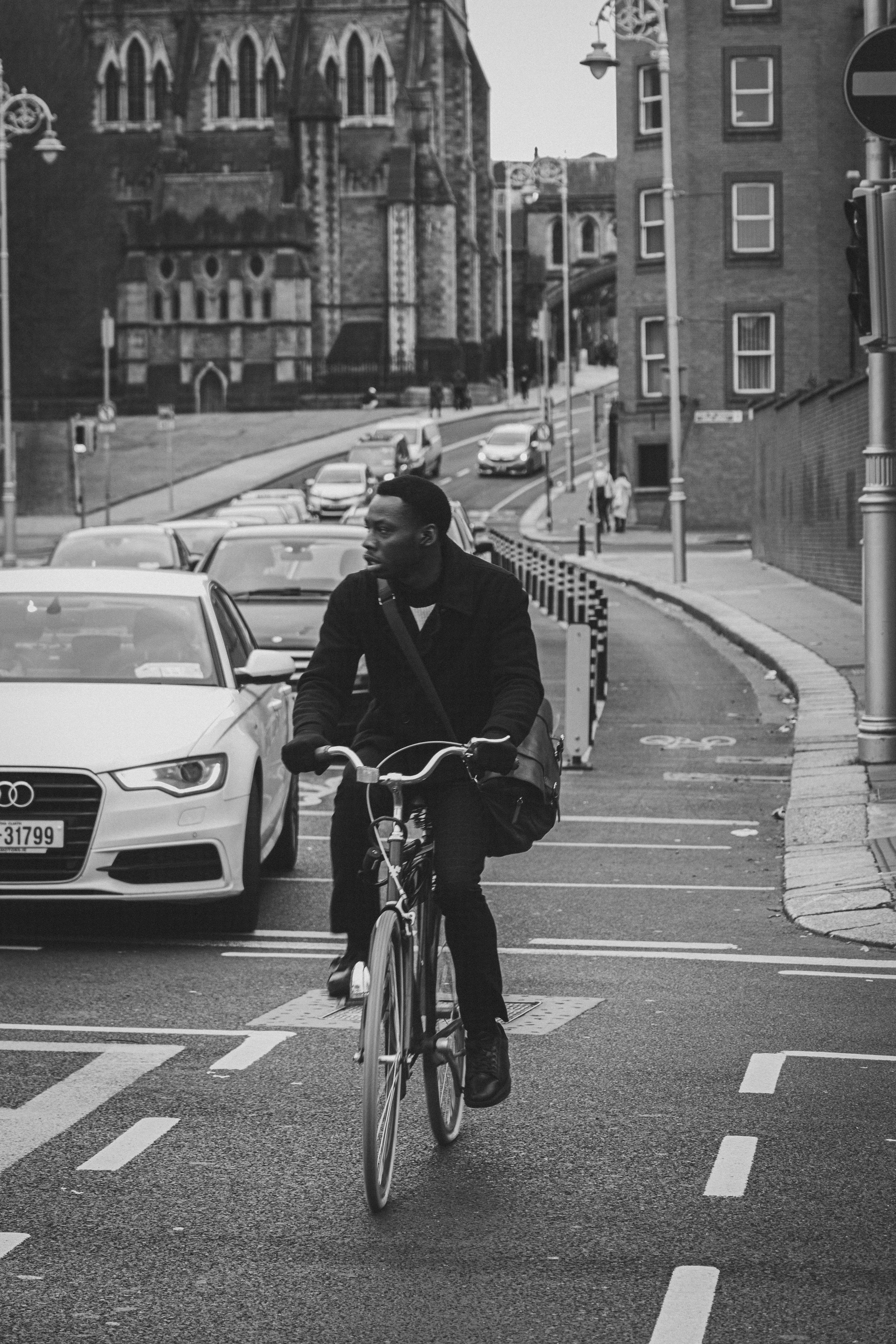 Black and white photo of a cyclist riding in urban traffic near historic architecture.