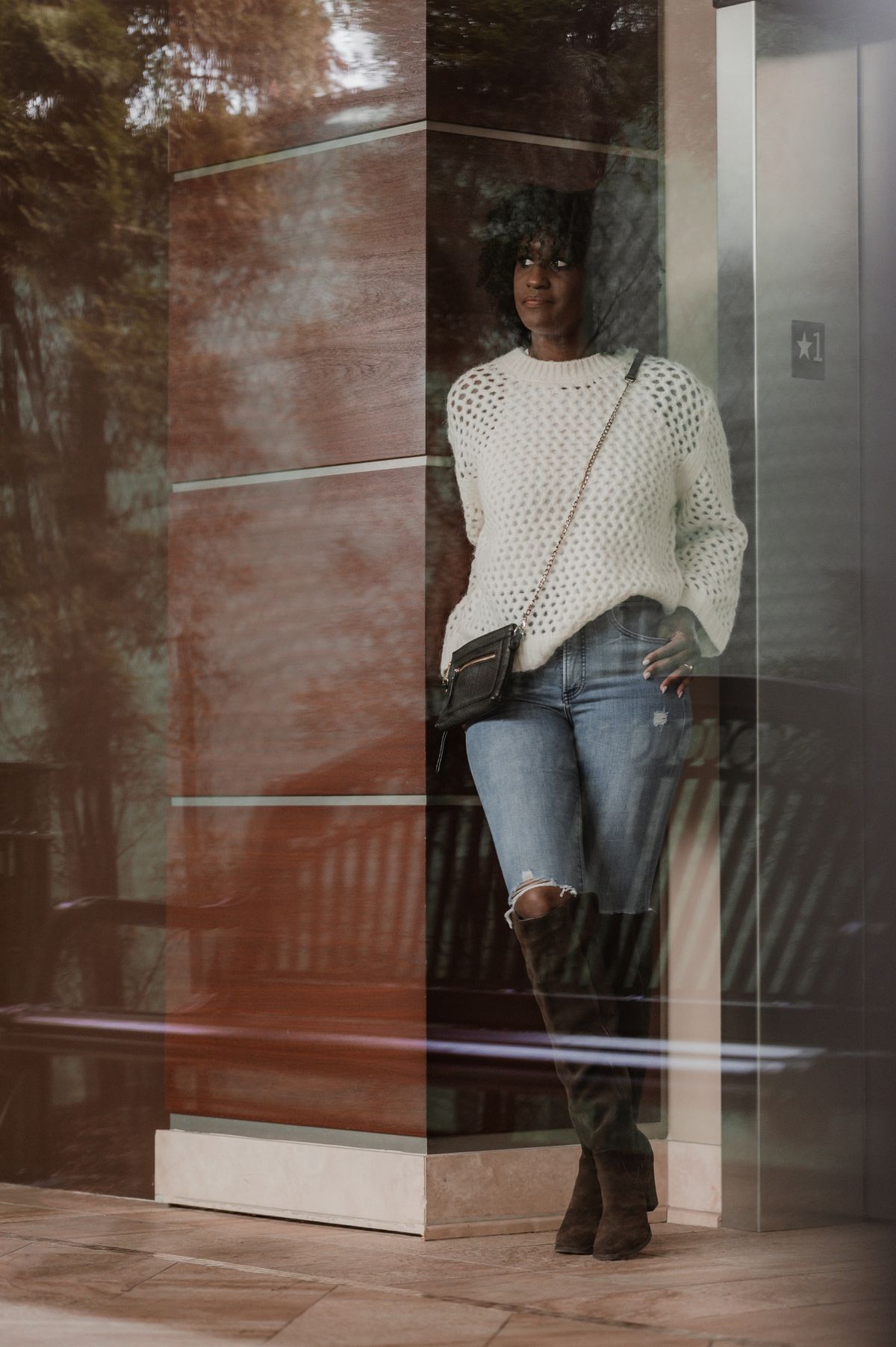 Fashionable woman in jeans and white sweater standing indoors against reflective glass surface.