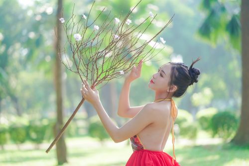 Free A beautiful woman in a red backless blouse holding a flowering twig in a lush green outdoor setting. Stock Photo